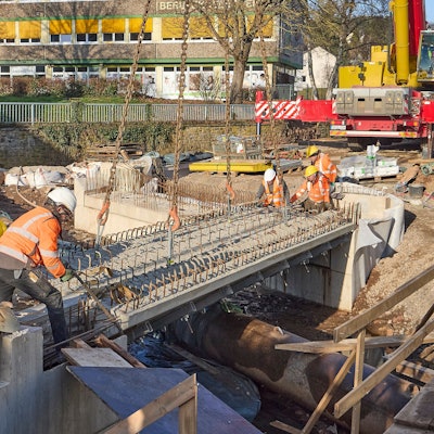 Fünf Männer tragen Warnjacken und Baustellenhelme. Sie bringen ein Bauteil einer Brücke in die exakte Position.