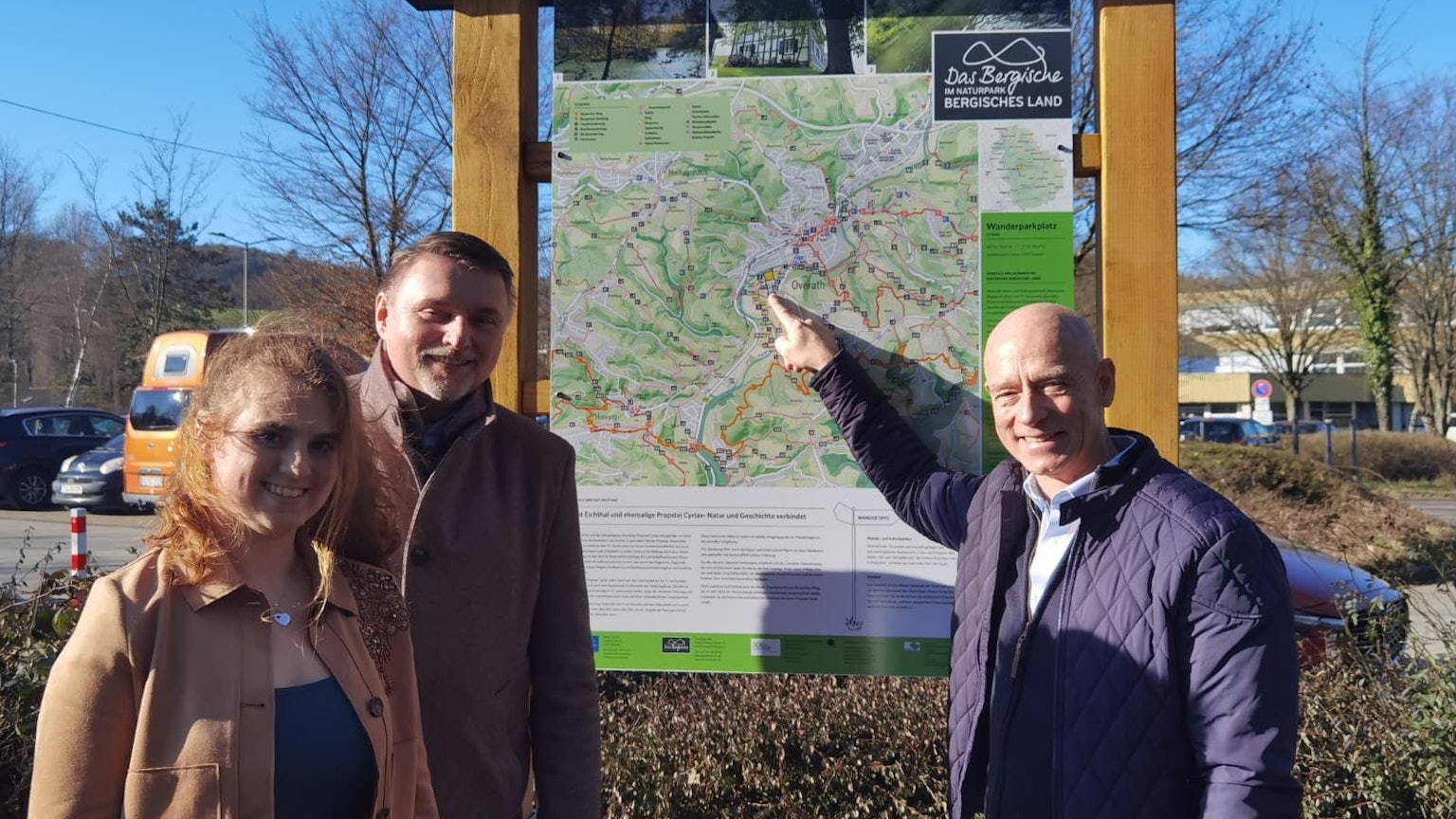 Eine Frau und zwei Männer stehen vor einer Wandertafel und lächeln.