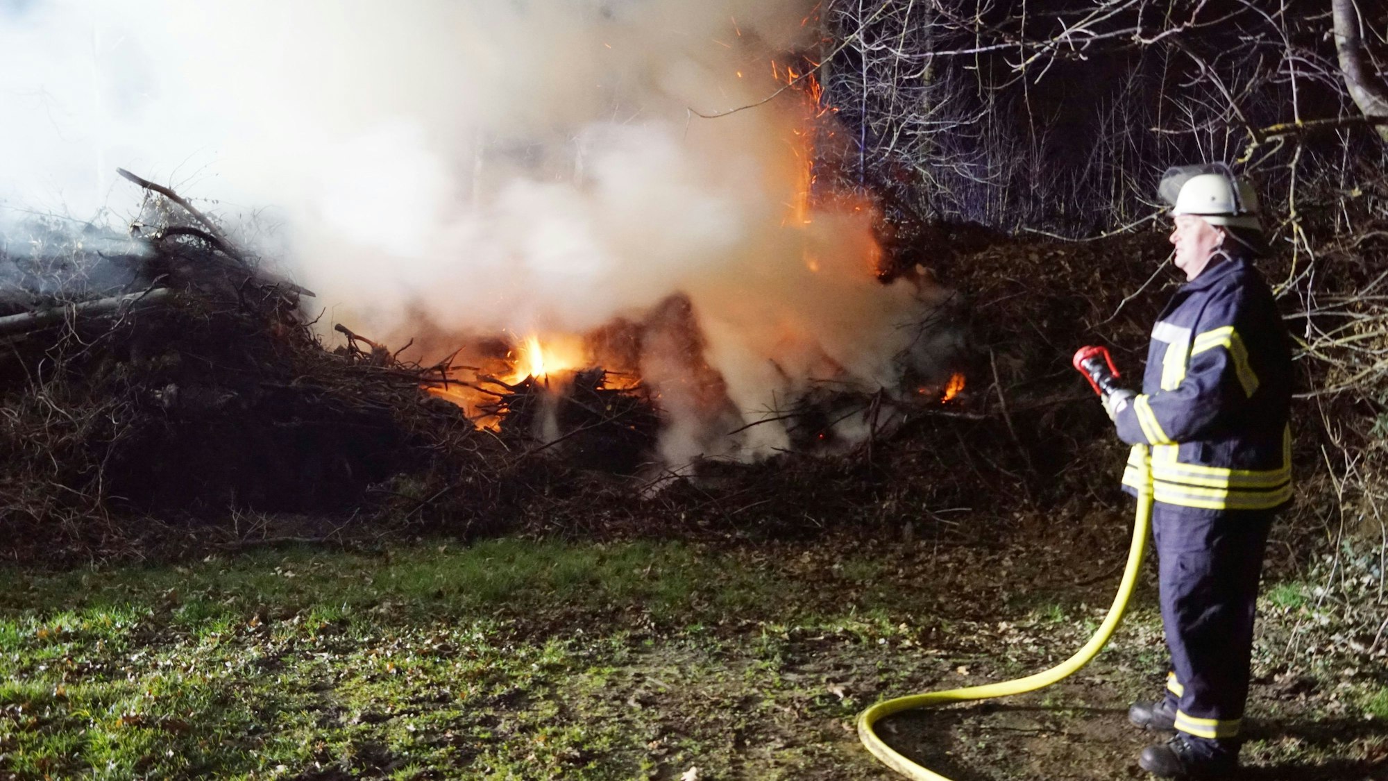Das Bild zeigt einen Feuerwehrmann, der einen Schlauch in den Händen hält. Im Hintergrund brennt Geäst.