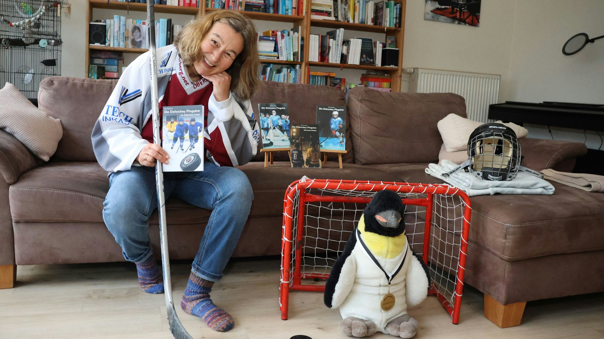 Elke Pfesdorf sitz auf einer Couch. In der Hand ein Buch und einen Hockeyschläger. Neben der Couch ist ein kleines Tor zusehen, in dem ein Pinguin steht.