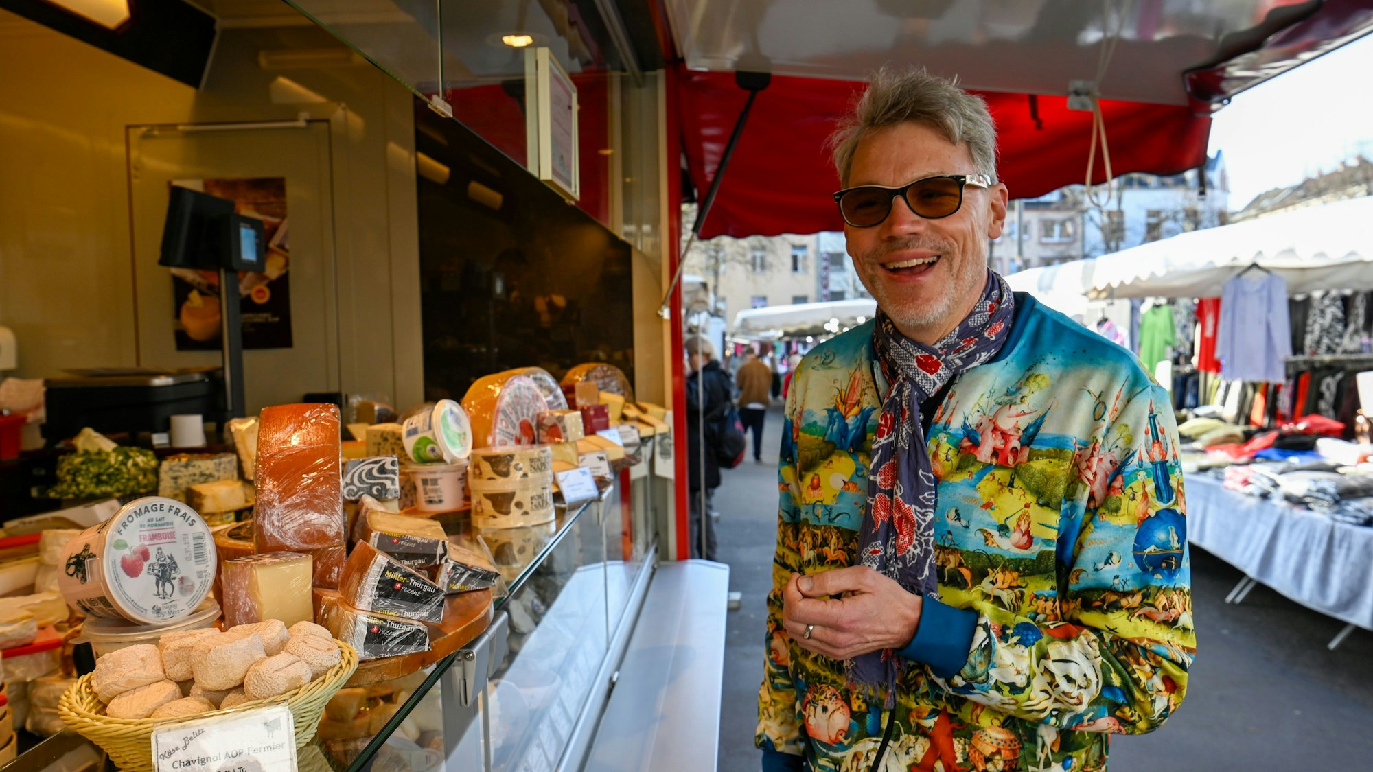 Ein Mann mit Sonnenbrille und bunter Jacke steht an einem Käsestand.