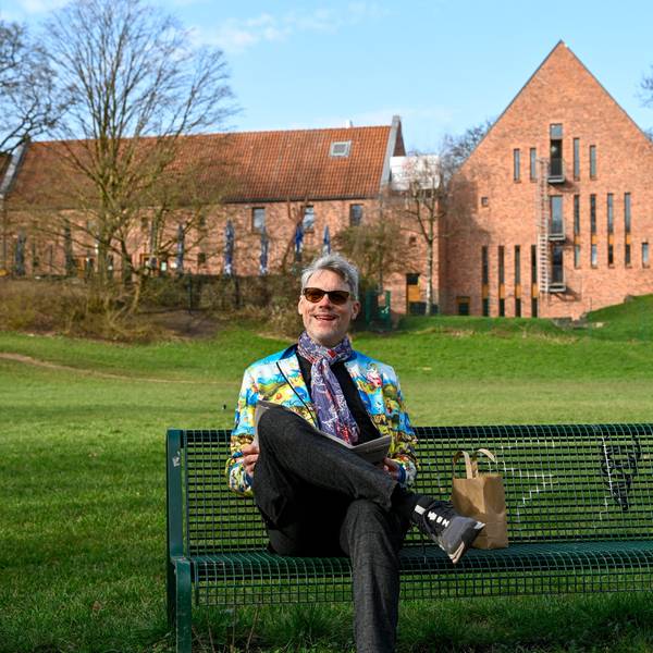 Ein Mann mit Sonnenbrille und bunter Jacke sitzt auf einer Bank, im Hintergrund ist der Altenberger Hof in Nippes zu sehen.