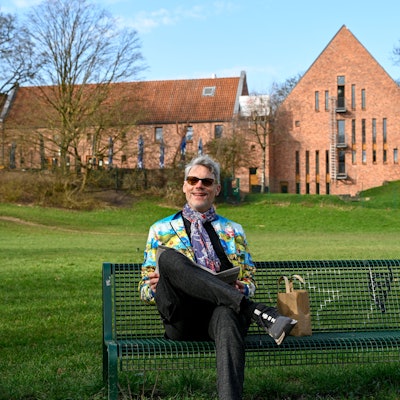 Ein Mann mit Sonnenbrille und bunter Jacke sitzt auf einer Bank, im Hintergrund ist der Altenberger Hof in Nippes zu sehen.