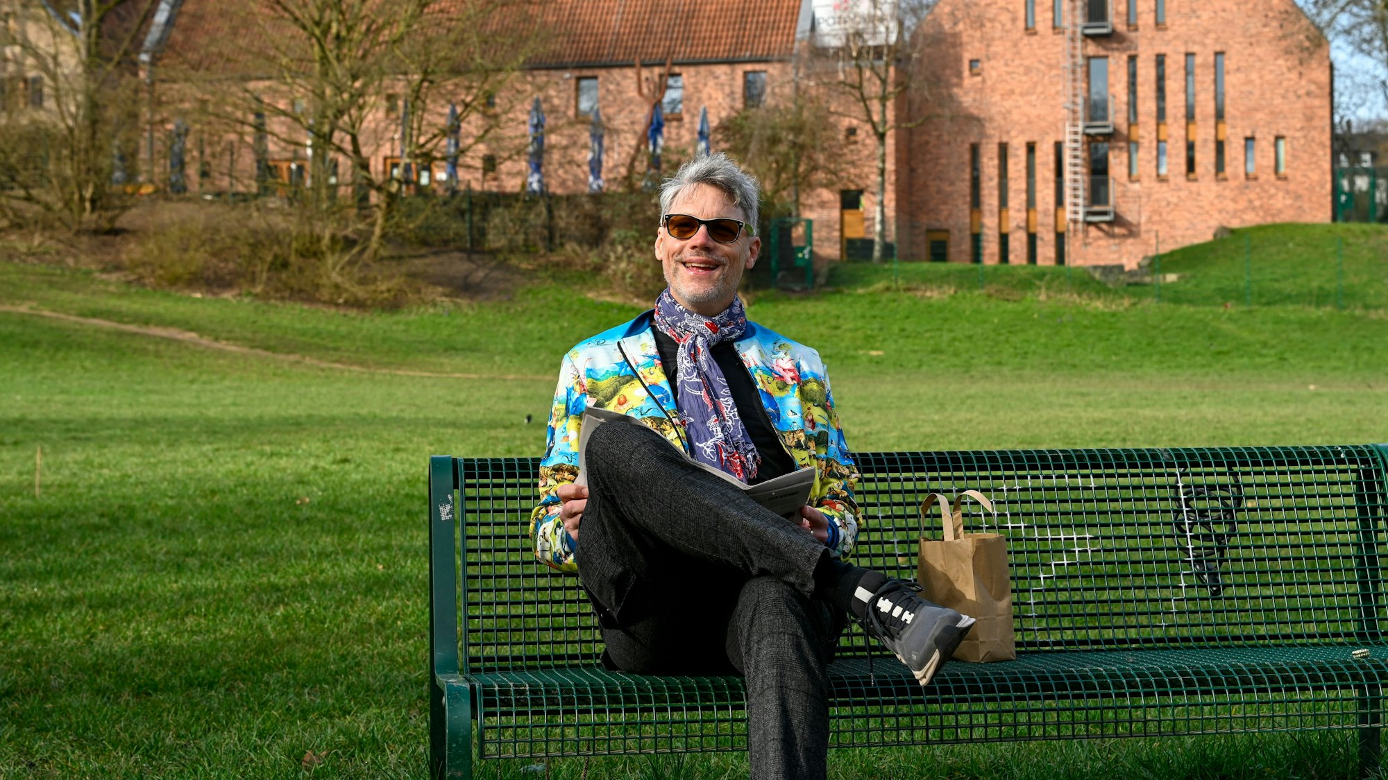 Ein Mann mit Sonnenbrille und bunter Jacke sitzt auf einer Bank, im Hintergrund ist der Altenberger Hof in Nippes zu sehen.