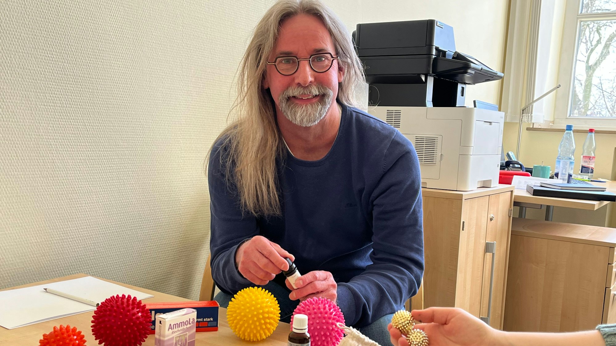 Ein Mann mit grauem langem Haar, Brille und Bart sitzt an einem Tisch. Neben ihm offenbar eine Patientin, man sieht nur ihre Hand, die ein stacheliges Kügelchen hält.