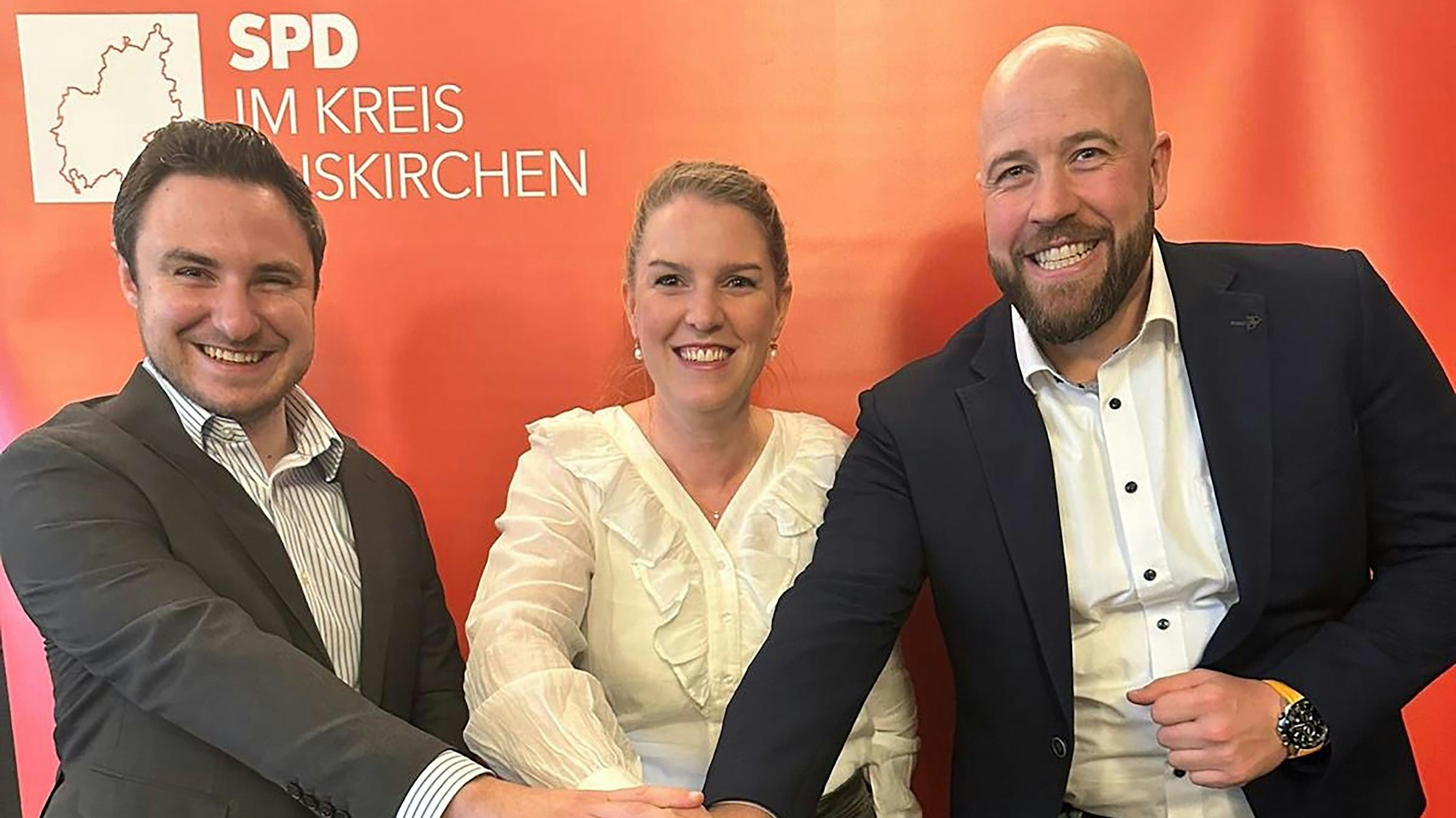 Die Obengenannten stehen vor einem SPD-Plakat und halten ihre rechten Hände aufeinander.