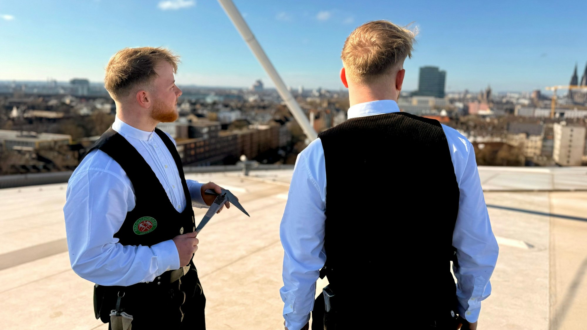 Noah und Noel Mohr stehen auf dem Dach der Lanxess-Arena und schauen auf die Skyline von Köln. Sie tragen ihre Arbeitskleidung, eine schwarze Weste und ein weißes Hemd darunter.