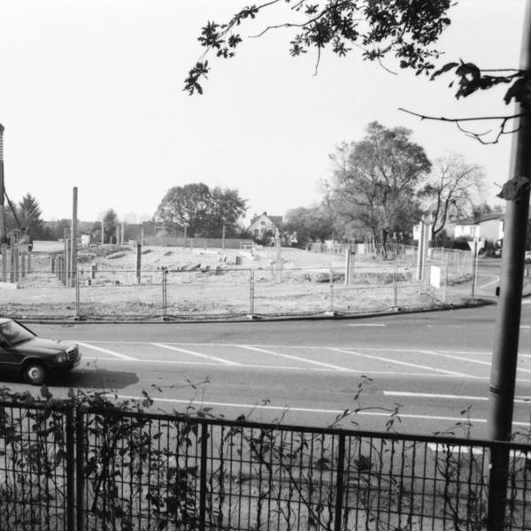 Man sieht ein altes schwarz-weiß Bild mit einem vorbeifahrenden Auto vor einer Baustelle.
