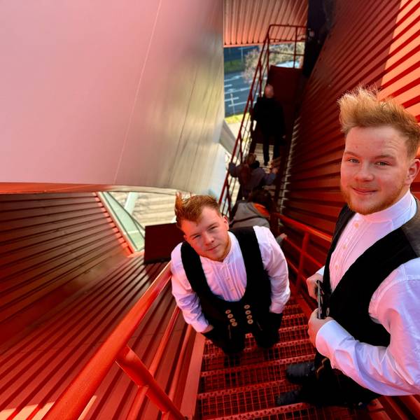 Noah und Noel Mohr stehen auf einer roten, schmalen Treppe, die auf das Dach der Lanxess-Arena führt. Sie tragen ihre Arbeitskleidung, eine schwarze Weste und ein weißes Hemd darunter.
