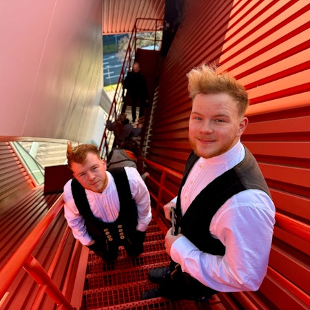 Noah und Noel Mohr stehen auf einer roten, schmalen Treppe, die auf das Dach der Lanxess-Arena führt. Sie tragen ihre Arbeitskleidung, eine schwarze Weste und ein weißes Hemd darunter.