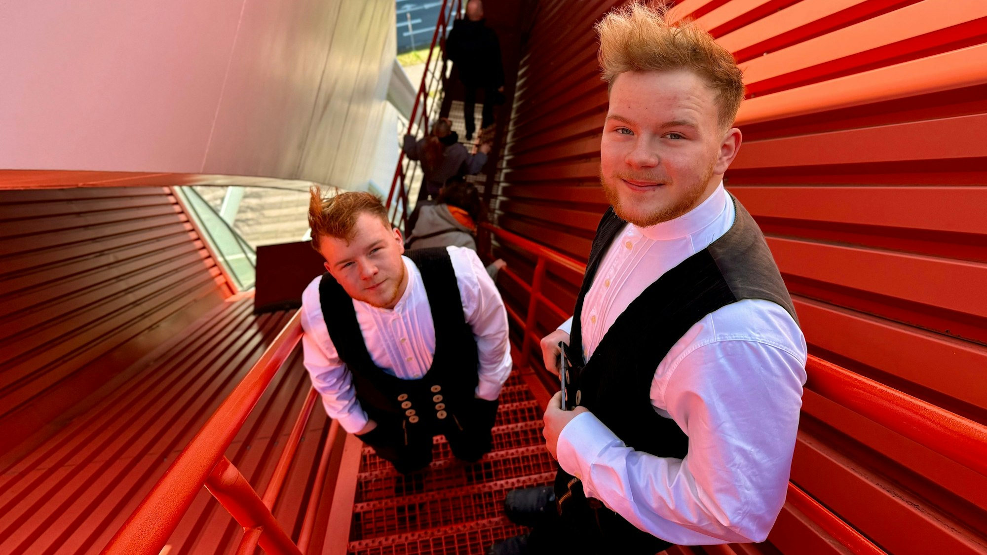 Noah und Noel Mohr stehen auf einer roten, schmalen Treppe, die auf das Dach der Lanxess-Arena führt. Sie tragen ihre Arbeitskleidung, eine schwarze Weste und ein weißes Hemd darunter.