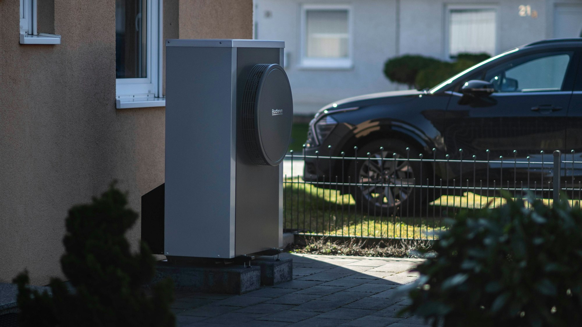 Vor einer Hausfassade steht eine Wärmepumpe. Im Bildhintergrund steht ein Auto in einer Einfahrt.