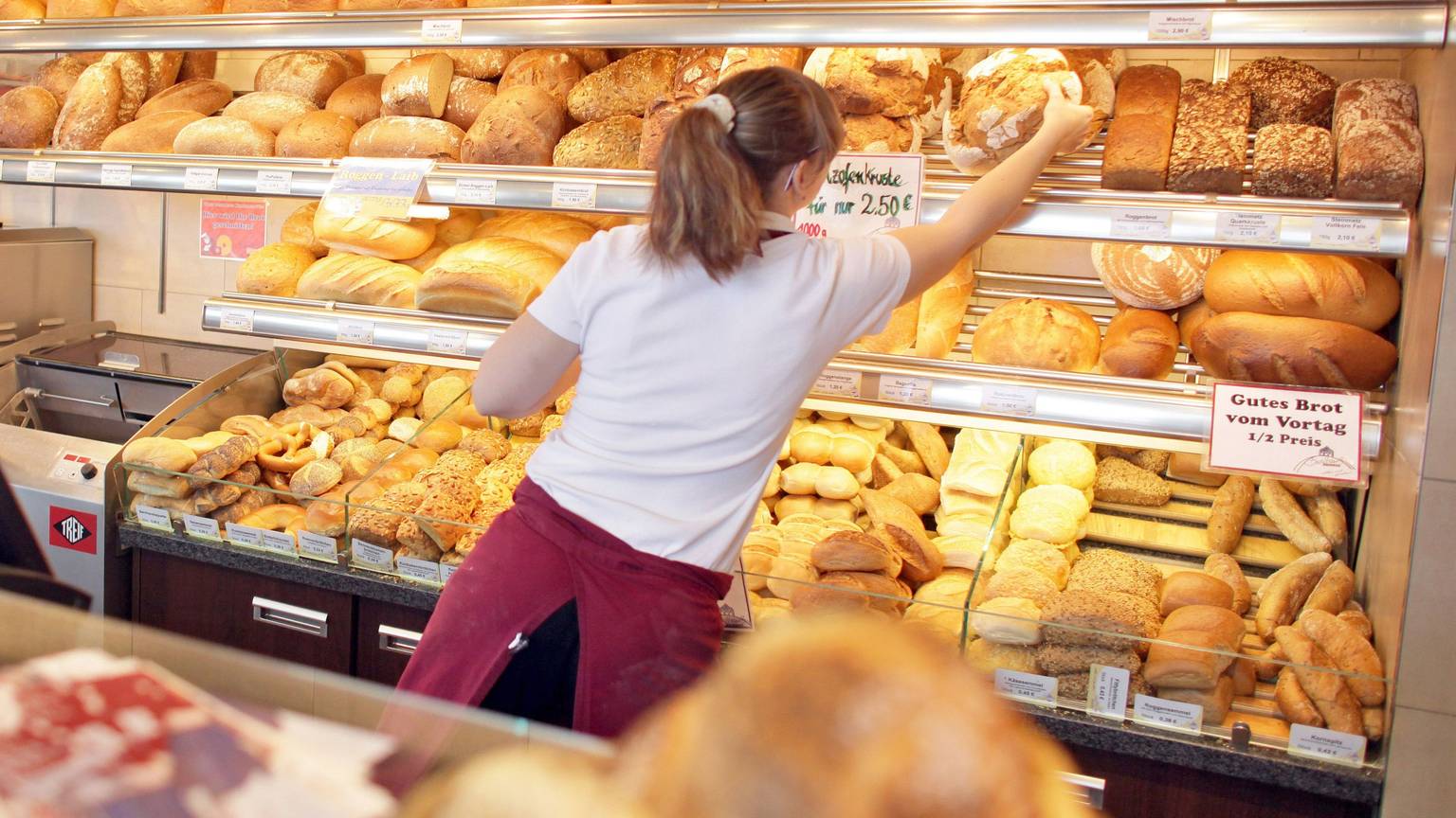 Eine Verkäuferin holt in einer Bäckerei ein Brot aus dem Regal.