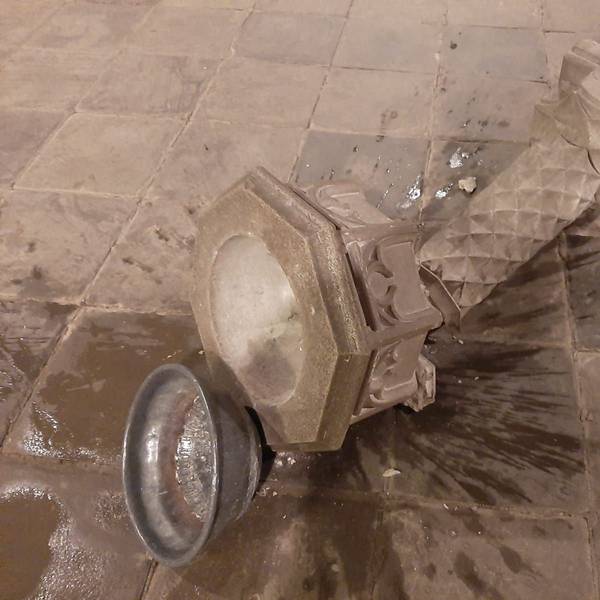 Eine Steinsäule mit Weihwasserbecken liegt zerbrochen am Boden, eine Metallschale daneben, Wasser ist ausgelaufen.