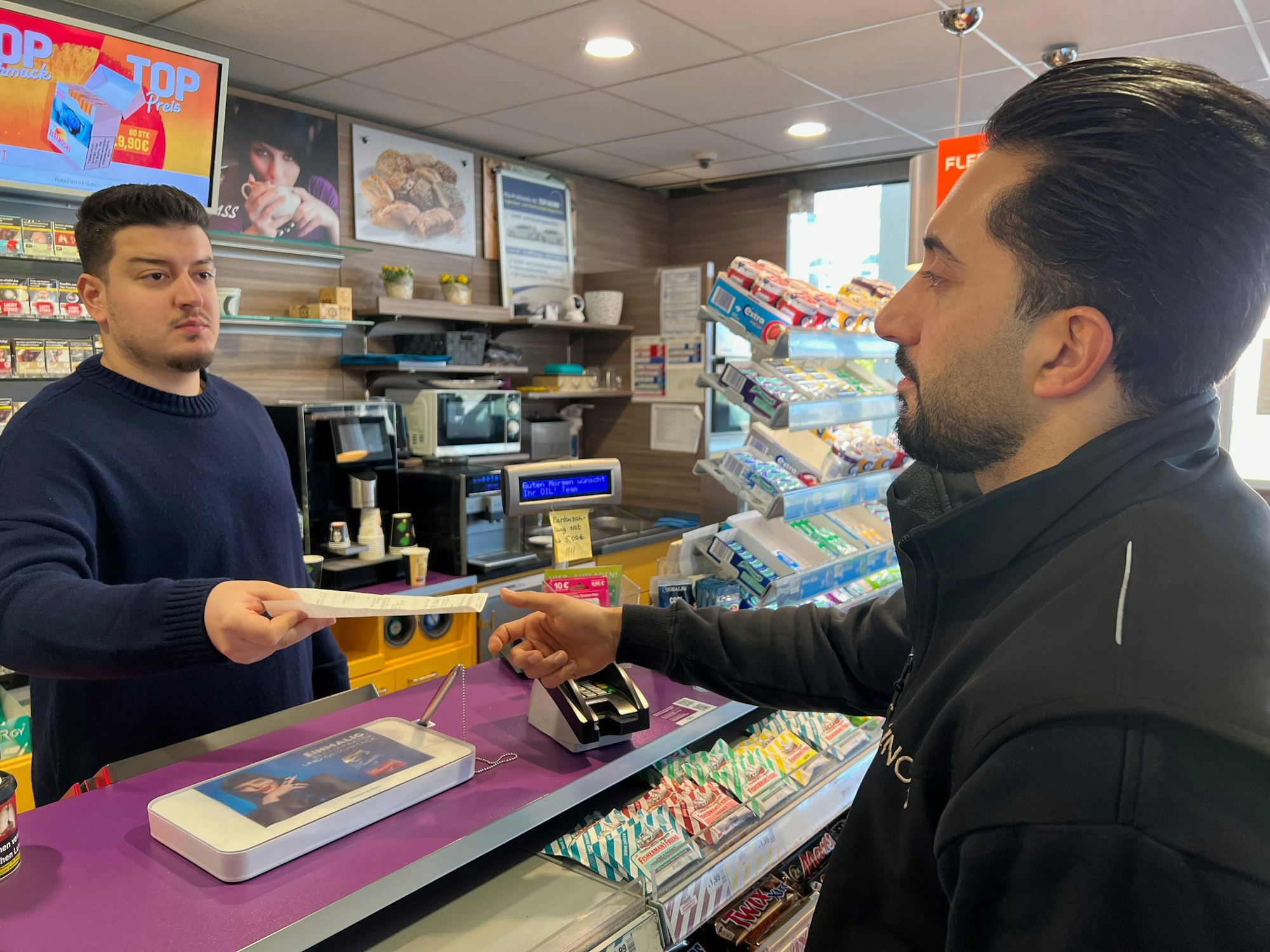 Das Bild zeigt zwei Männer an der Kasse einer Tankstelle.