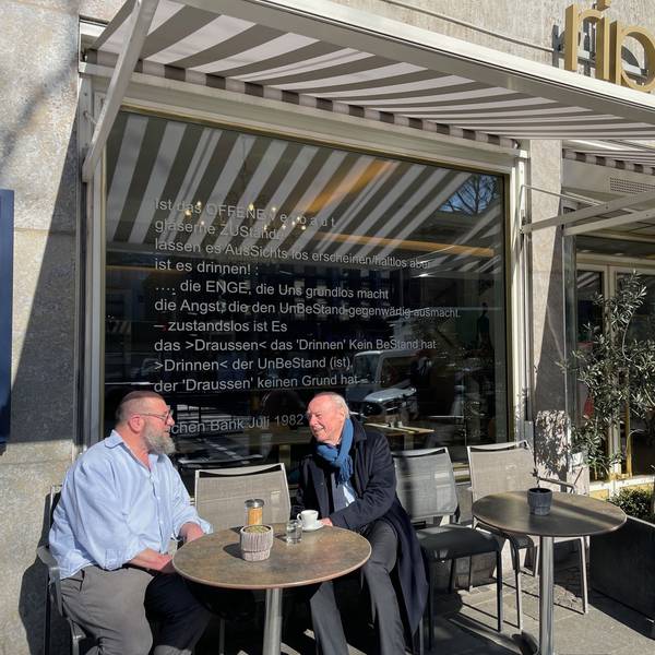Ein Mann mit Bart und ein Senior in Schal und Mantel sitzen an einem Tisch draußen vor einem Restaurant.