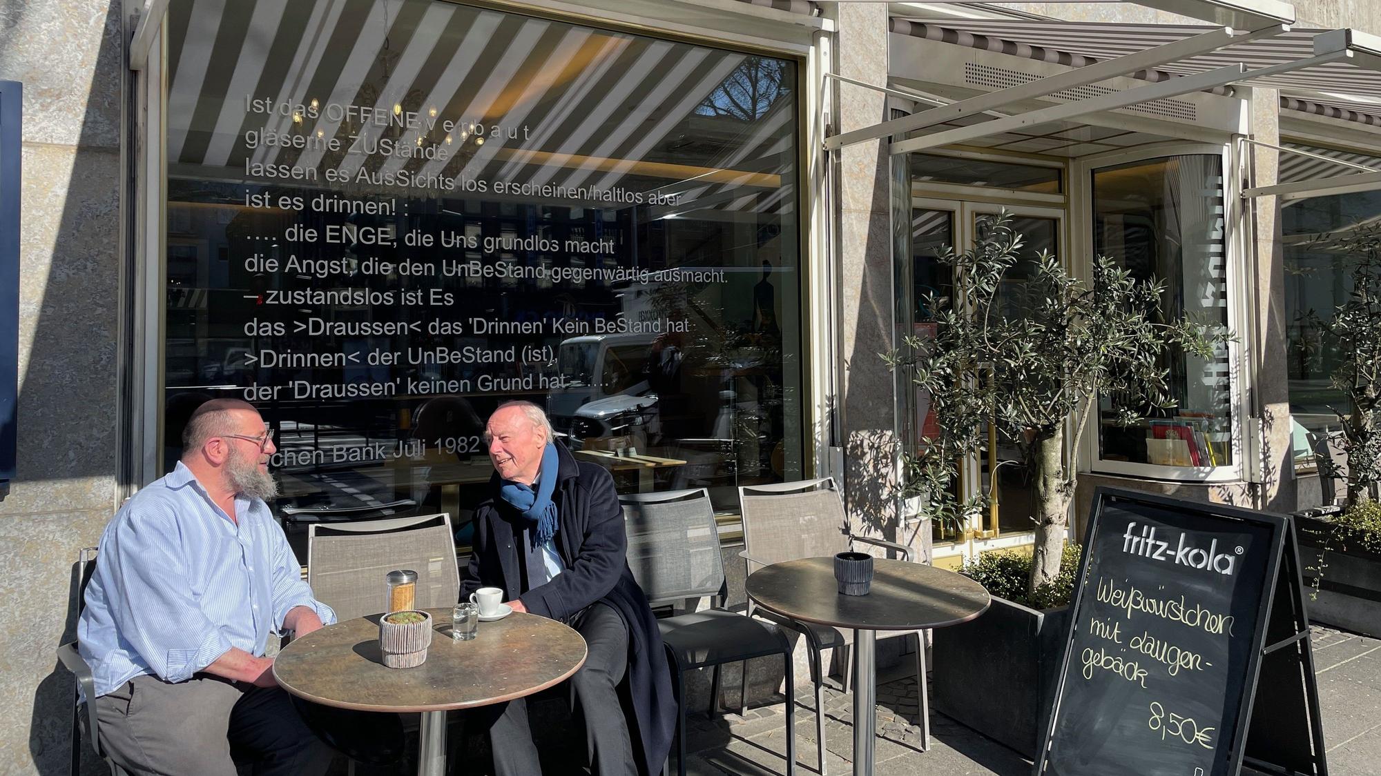 Ein Mann mit Bart und ein Senior in Schal und Mantel sitzen an einem Tisch draußen vor einem Restaurant.