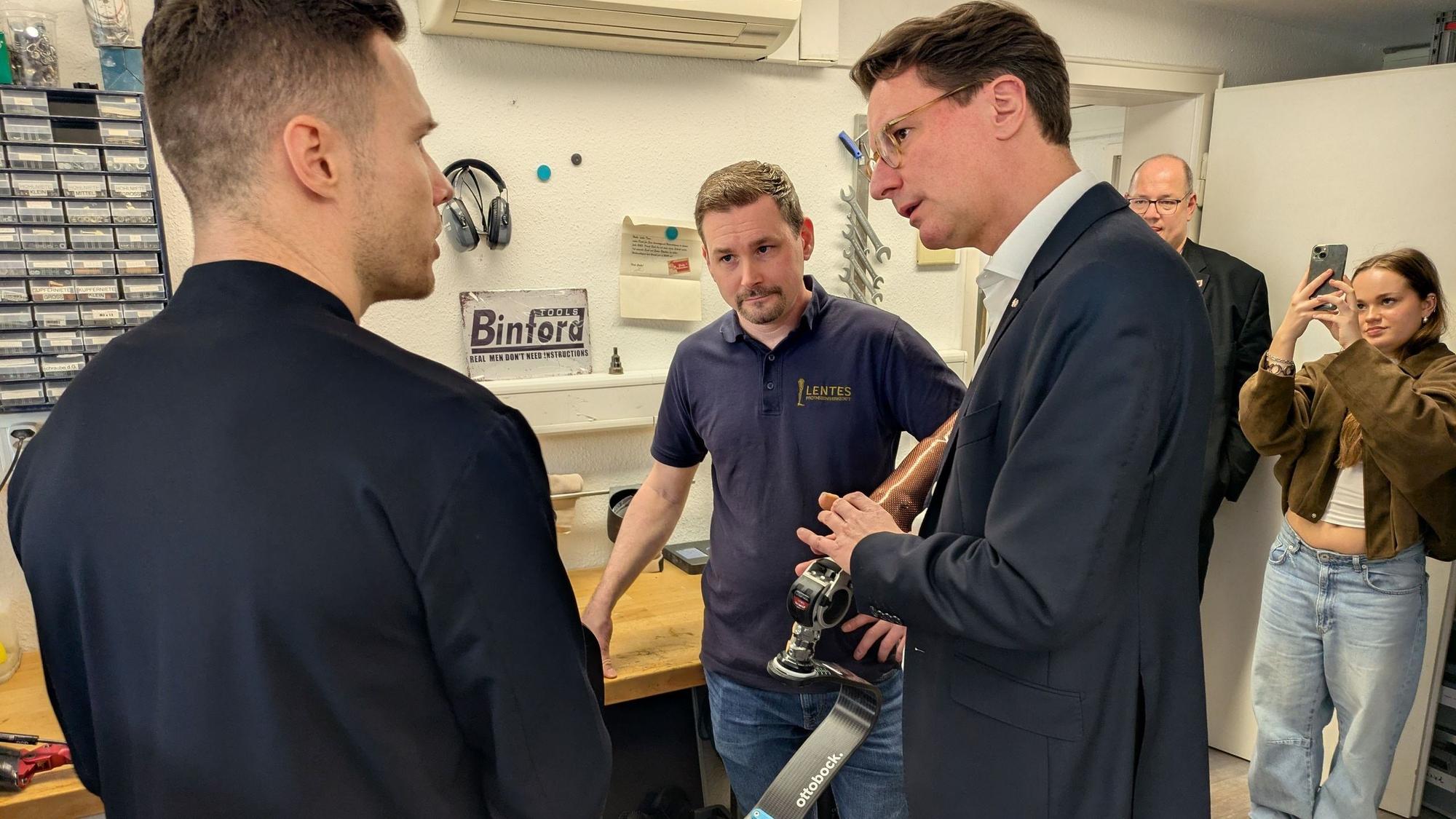 Ministerpräsident Hendrik Wüst lässt sich von Tim von Hagen (M.) und Markus Rehm die Besonderheiten von Rehms Wettkampfprothese erläutern.