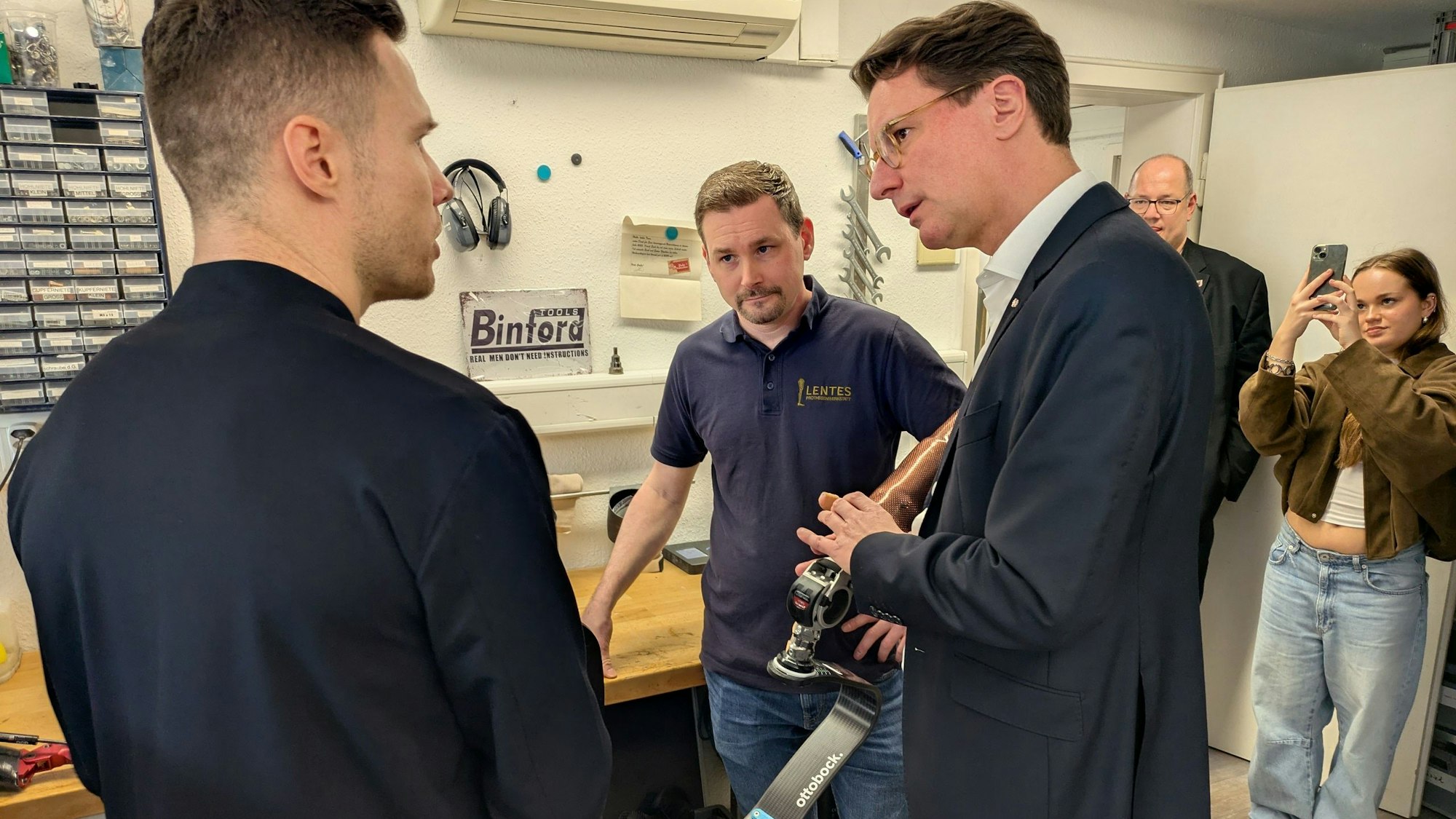 Ministerpräsident Hendrik Wüst lässt sich von Tim von Hagen (M.) und Markus Rehm die Besonderheiten von Rehms Wettkampfprothese erläutern.