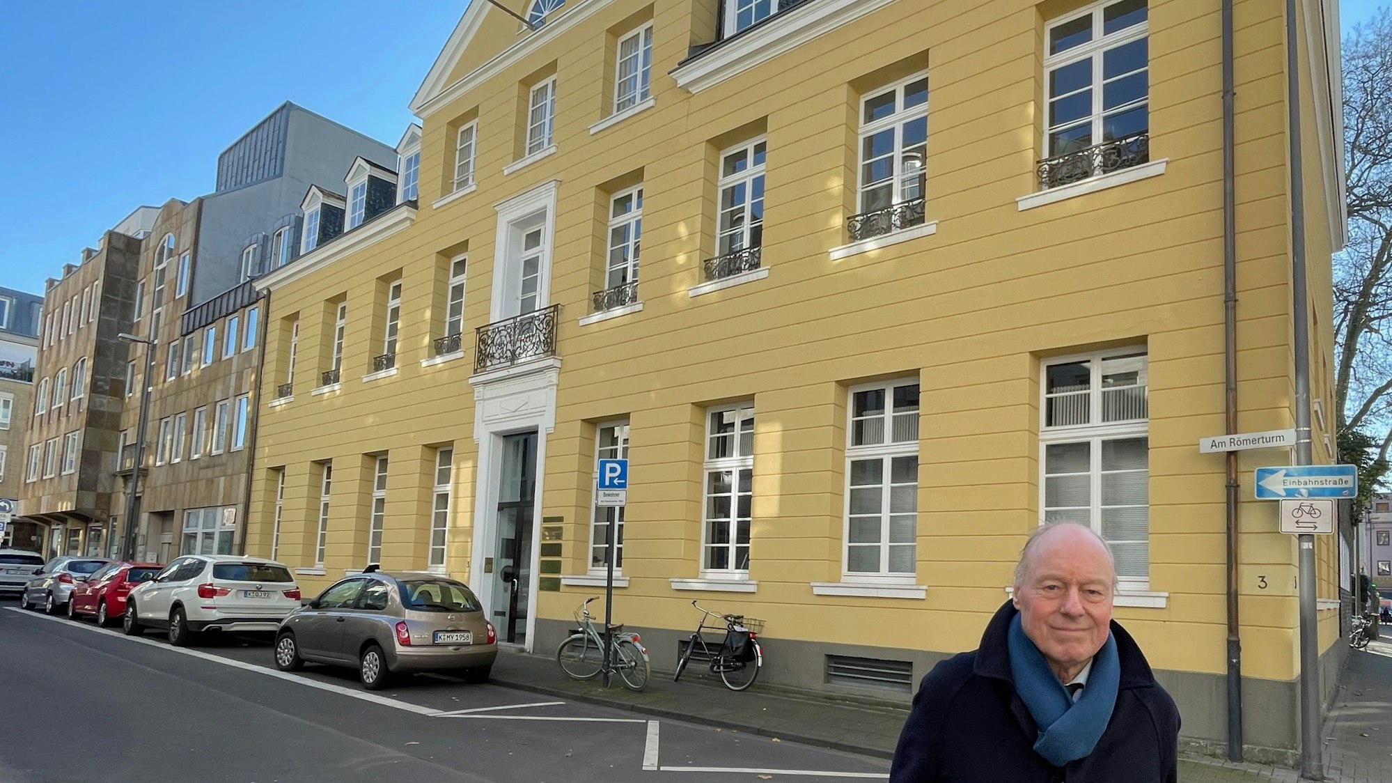 Vor einem großen Haus mit gelber Fassade im klassizistischen Stil steht ein Mann in Mantel und Schal.