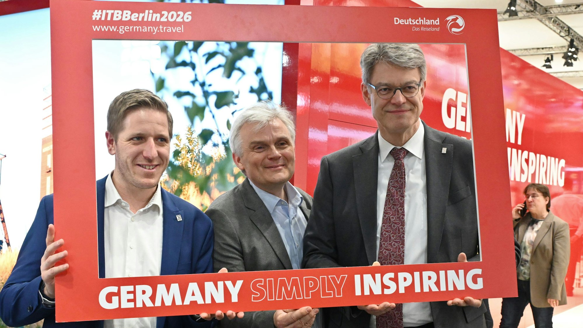 Bundesverkehrsminister Patrick Schnieder (v.r.), Wolfgang Reh und Markus Ramers blicken auf der ITB Berlin durch einen roten Fotorahmen mit der Aufschrift „Germany simply inspiring“