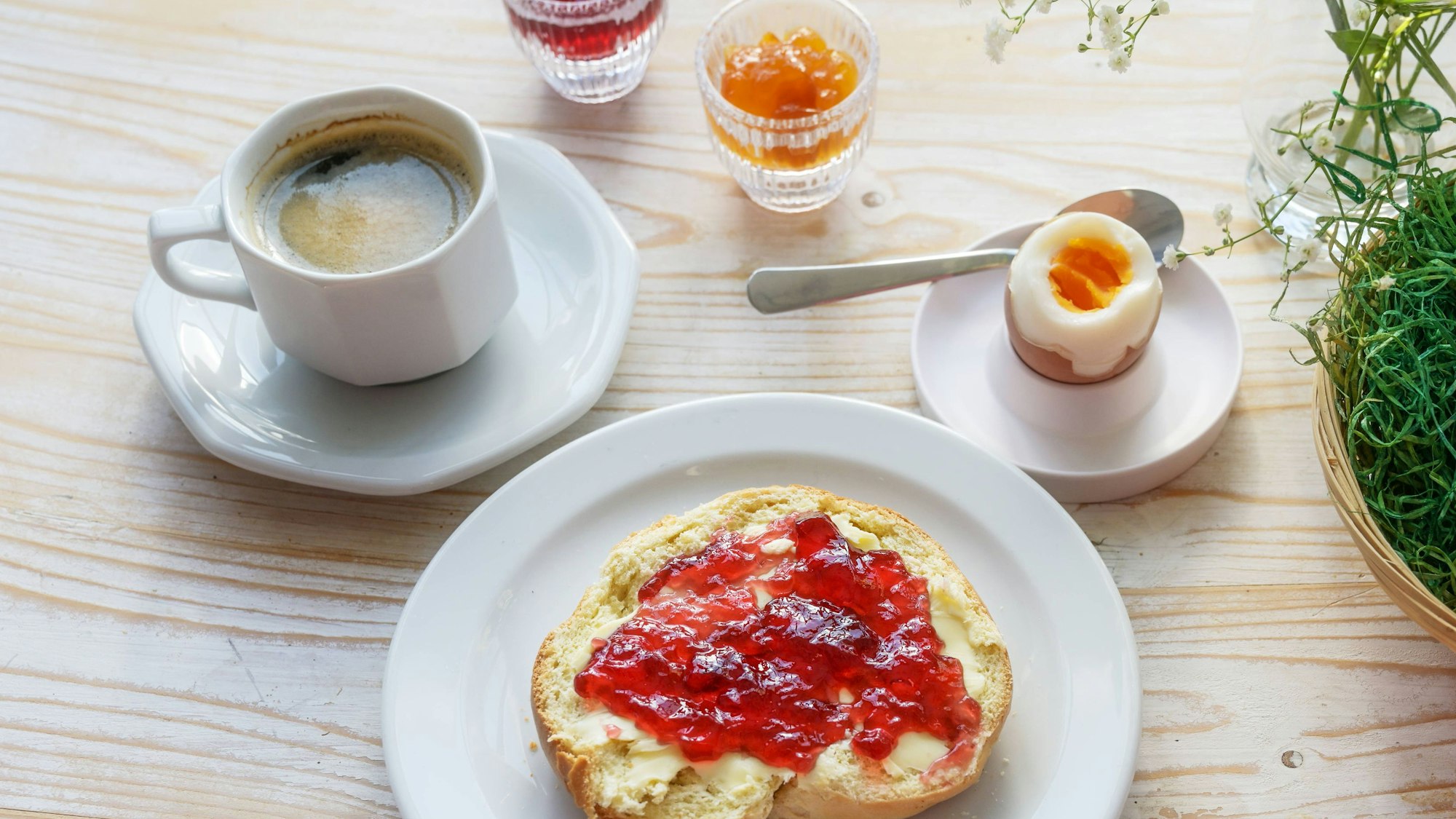 Osterfrühstück, Brötchen oder Sandwich mit roter Kirschmarmelade, gekochtes Ei, Kaffee und ein Korb mit braunen Eiern auf einem Holztisch.