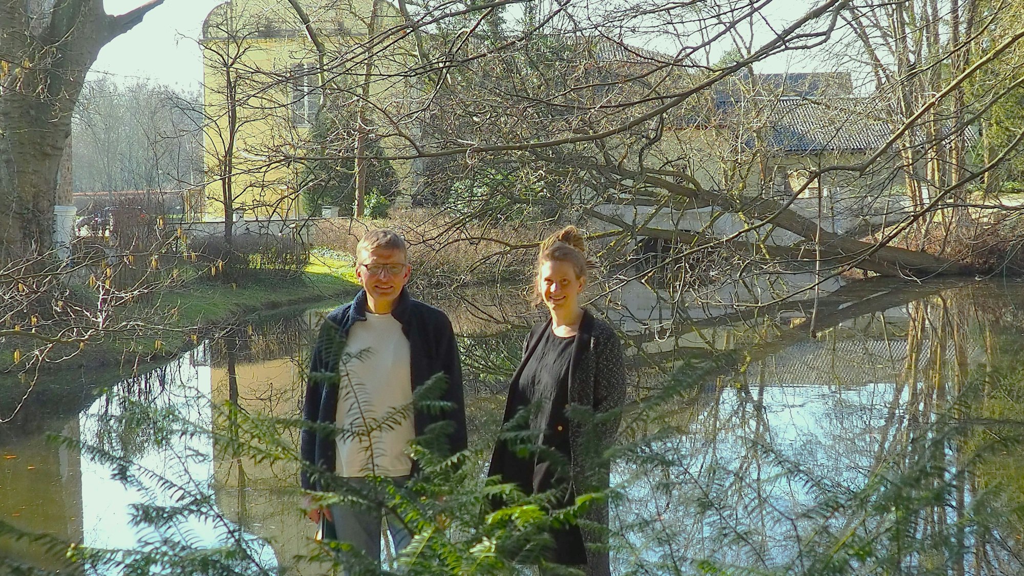 Dorothee Lindenbaum und Stefan Bergner (l.) stehen am Wassgeraben, in dem sich die Burg Flamersheim spiegelt.