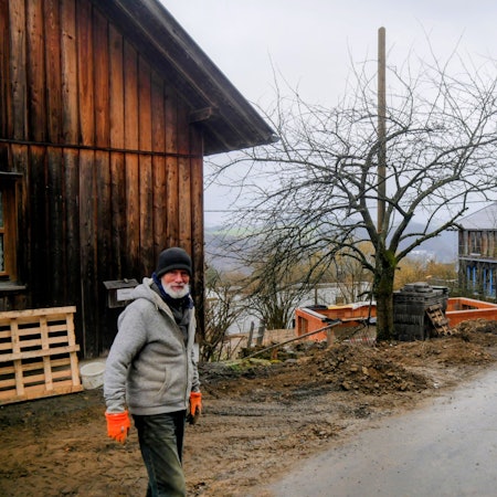 Ein Mann steht mit Mütze und Handschuhen auf der Straße. Im Hintergrund sind die Häuser, dass eine noch im Rohbau, zu sehen.