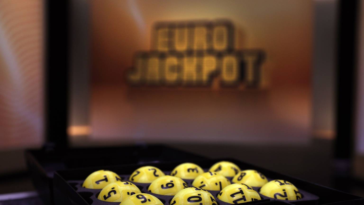 Das Bild zeigt mehrere gelbe Lotteriekugeln mit schwarzen Nummern, die ordentlich in einem schwarzen Tablett angeordnet sind. Im unscharfen Hintergrund ist das beleuchtete Eurojackpot-Logo zu erkennen.