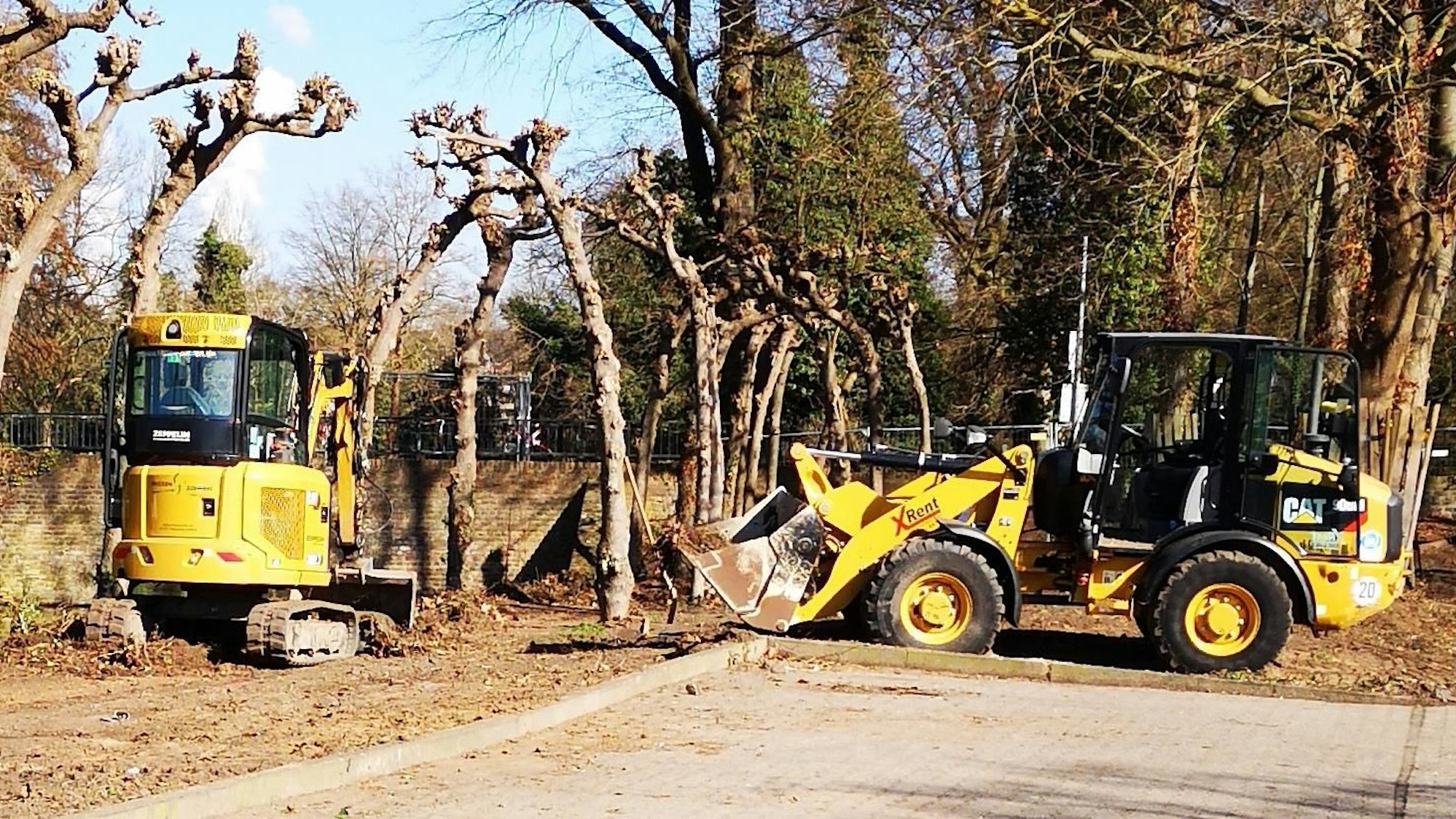 Ein Radlader und ein kleiner Bagger stehen nah an Bäumen.