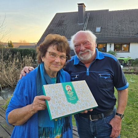 Ein älteres Ehepaar steht vor seinem Haus, sie hält eine Schachtel.