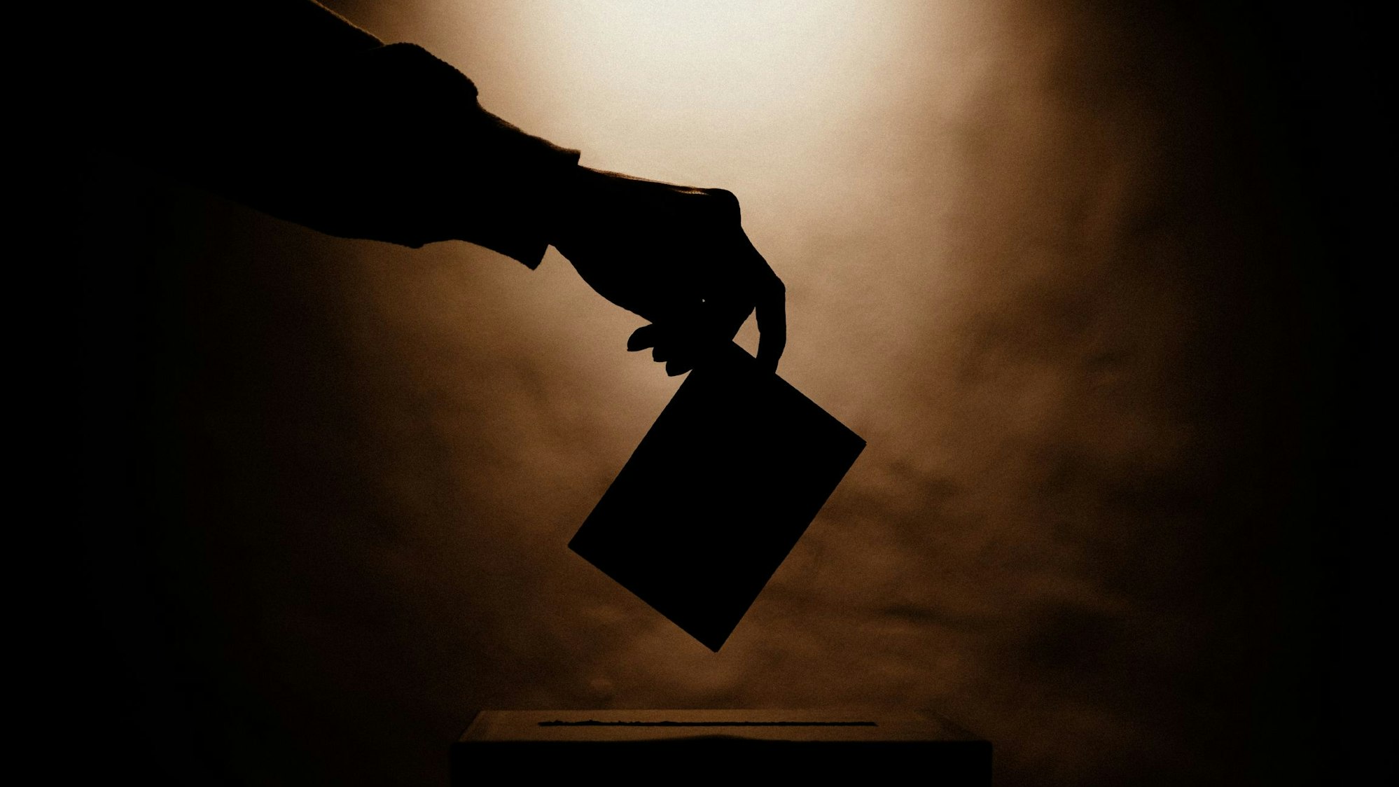 Eine Hand, die einen Wahlumschlag in eine Urne wirft.