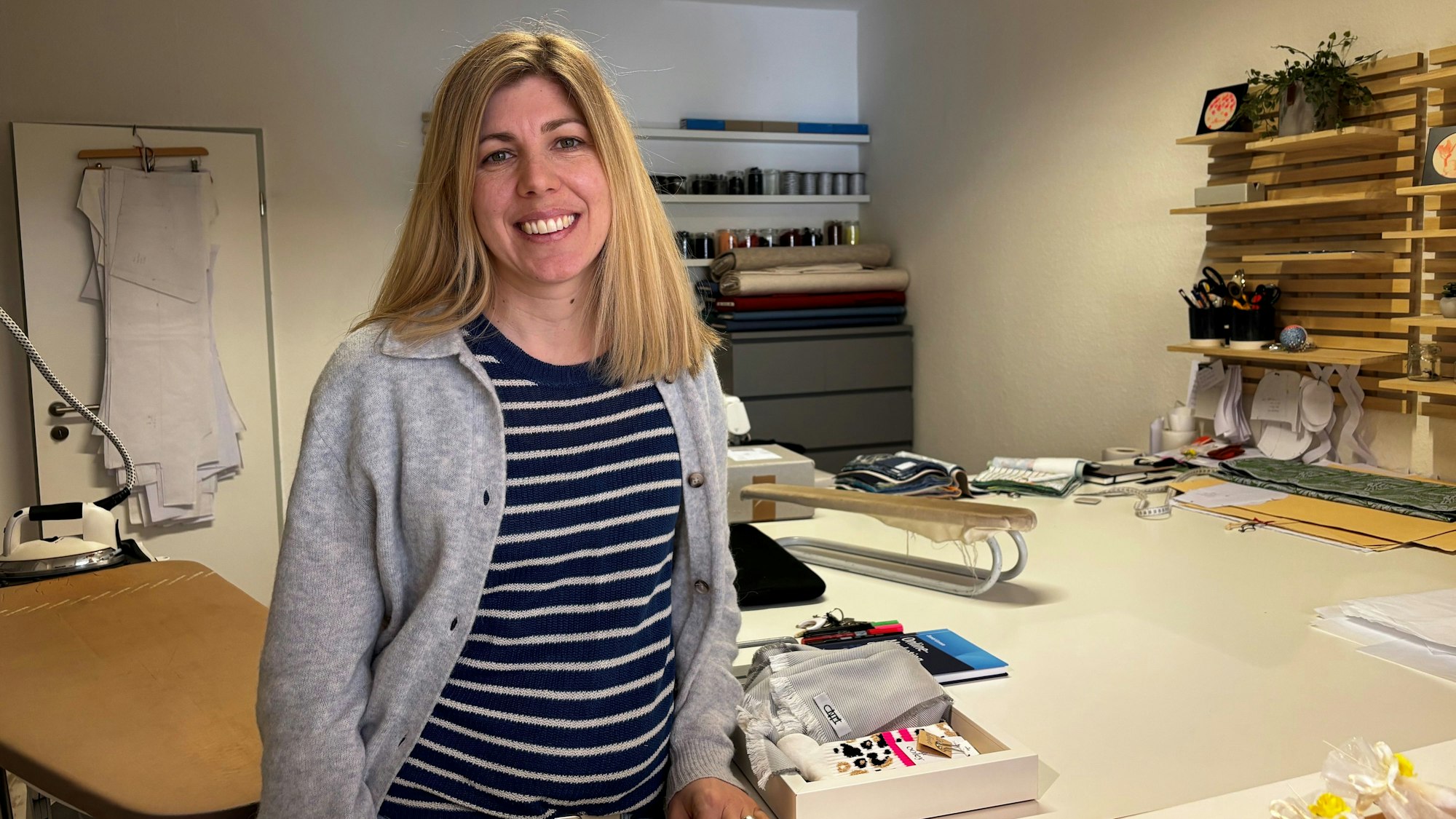 Carina Molitor steht in ihrem Atelier und blickt lachend in die Kamera. Im Hintergrund sieht man ein Bügelbrett, Schnittmuster und Stoffproben.