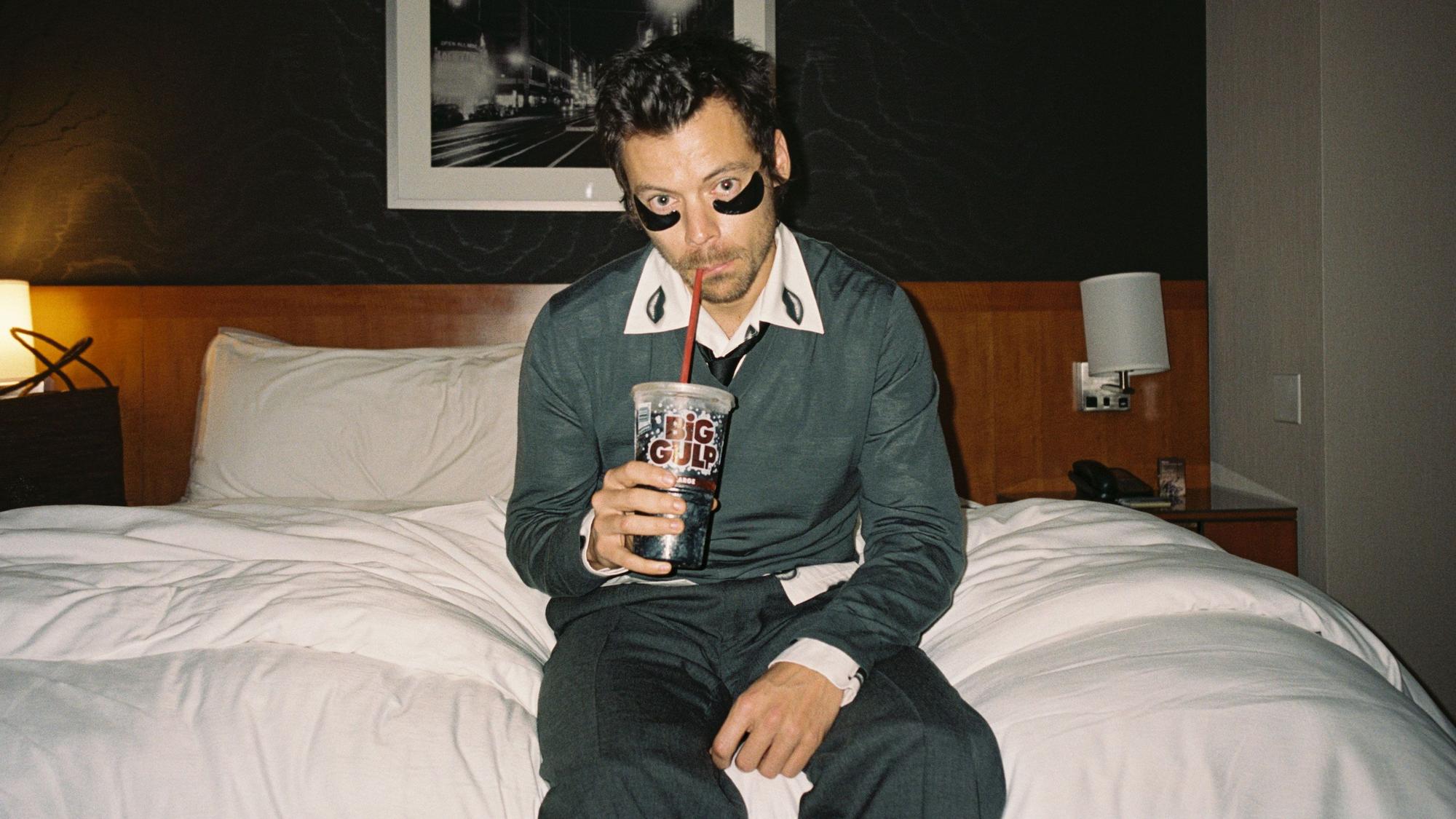 Harry Styles sitzt auf einem Hotelbett. Er trägt schwarze Augenpads gegen Tränensäcke und schlürft mit dem Strohhalm aus einem Plastikbecher.