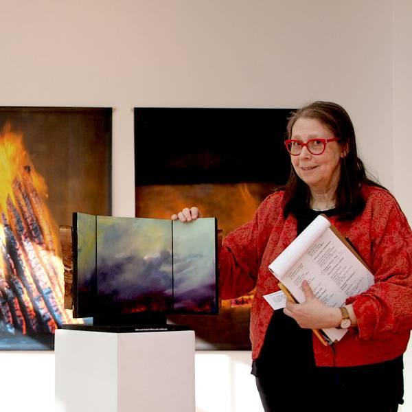 Die Kuratorin Rena Voss steht im Kunstforum Gemünd in der Ausstellung an einem Triptychon. Im Hintergrund sind Fotos mit Feuer zu sehen.
