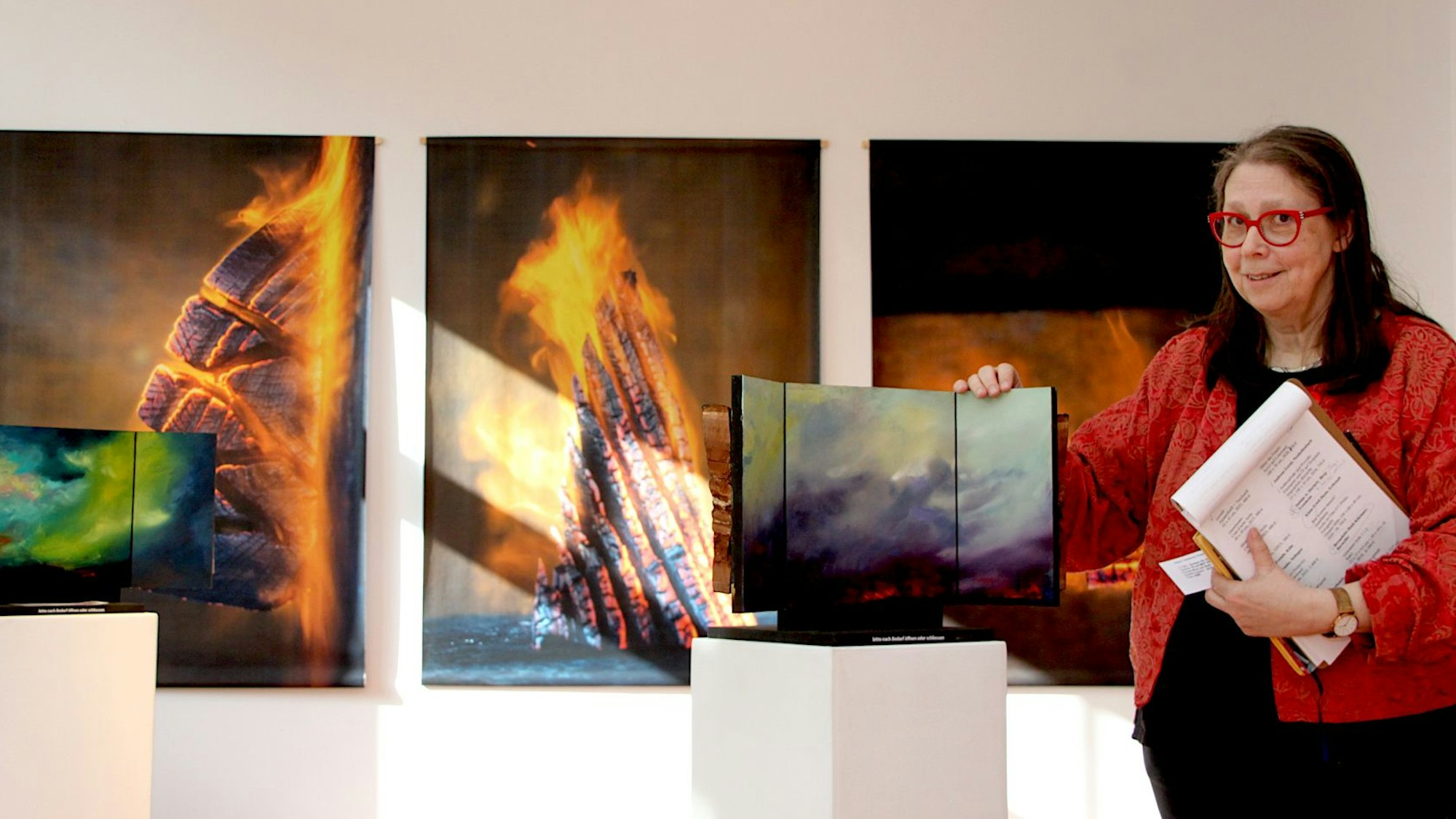 Die Kuratorin Rena Voss steht im Kunstforum Gemünd in der Ausstellung an einem Triptychon. Im Hintergrund sind Fotos mit Feuer zu sehen.