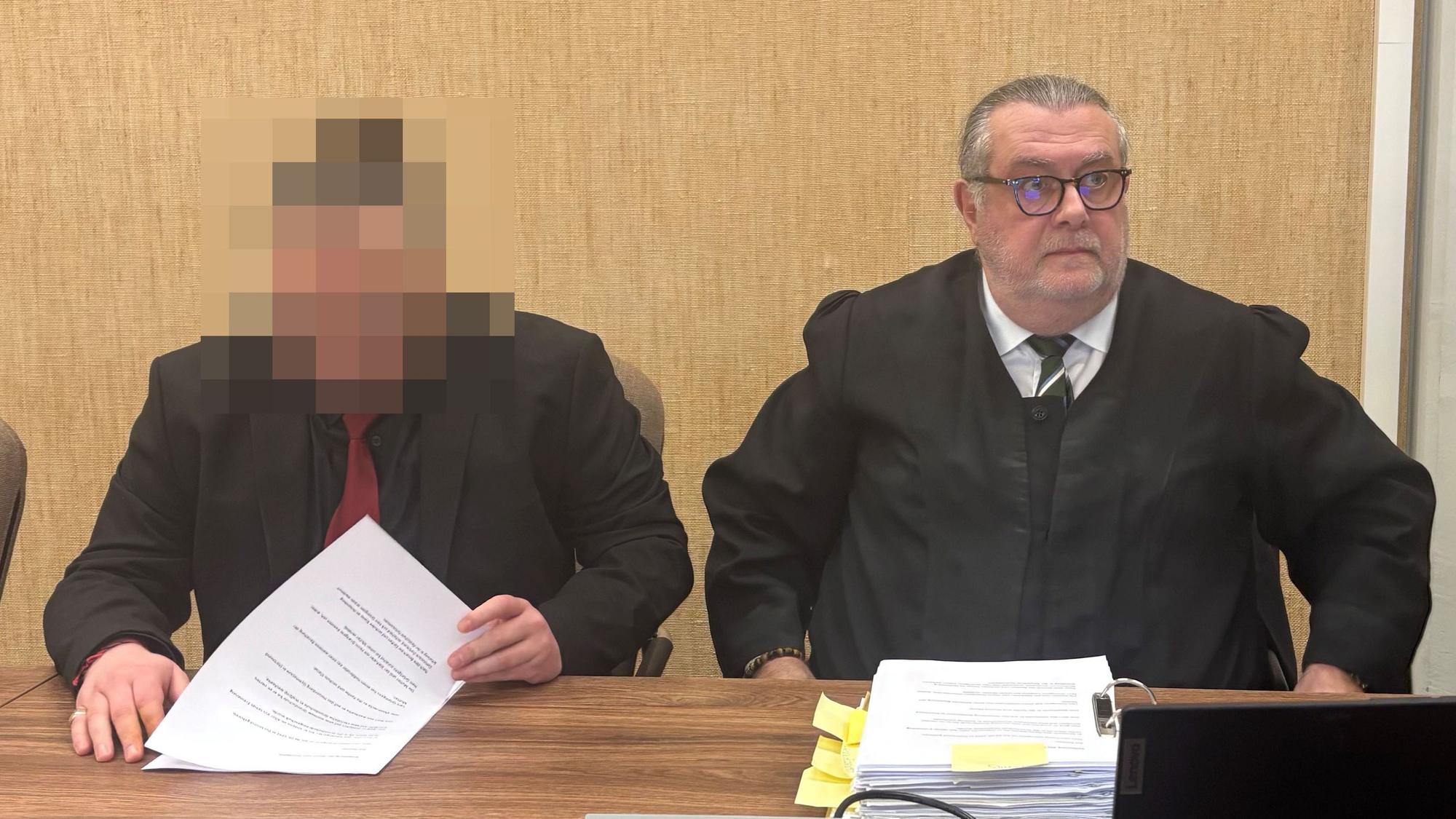 Der Angeklagte sitzt im schwarzen Anzug mit roter Krawatte neben seinem Verteidiger Thomas Düber.