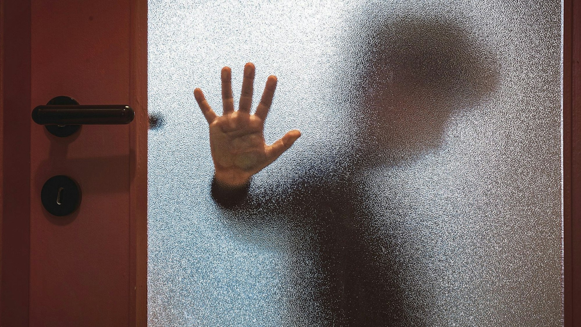 Ein Junge hält seine Hand abwehrend gegen eine Glasscheibe in der Tür, hinter der er sich versteckt hat.