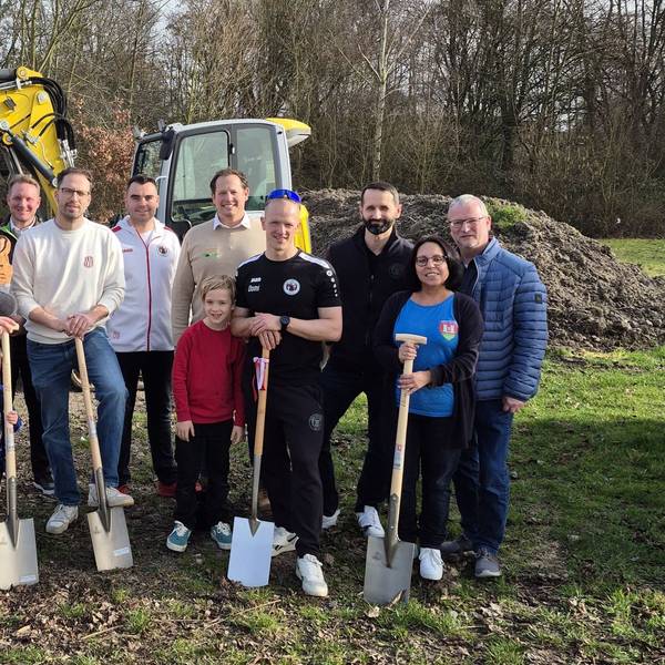 Männer, Frauen und Kinder stehen auf der Baustelle und halten teilweise Spaten in der Hand.
