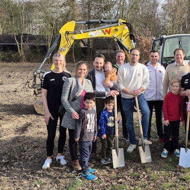 Männer, Frauen und Kinder stehen auf der Baustelle und halten teilweise Spaten in der Hand.