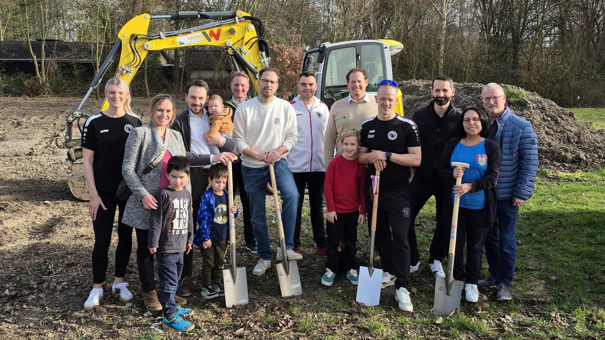 Männer, Frauen und Kinder stehen auf der Baustelle und halten teilweise Spaten in der Hand.