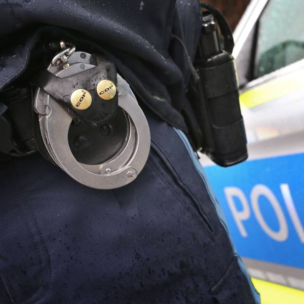 Ein Polizist mit Handschellen und einer Pistole am Gürtel steht vor einem Streifenwagen.