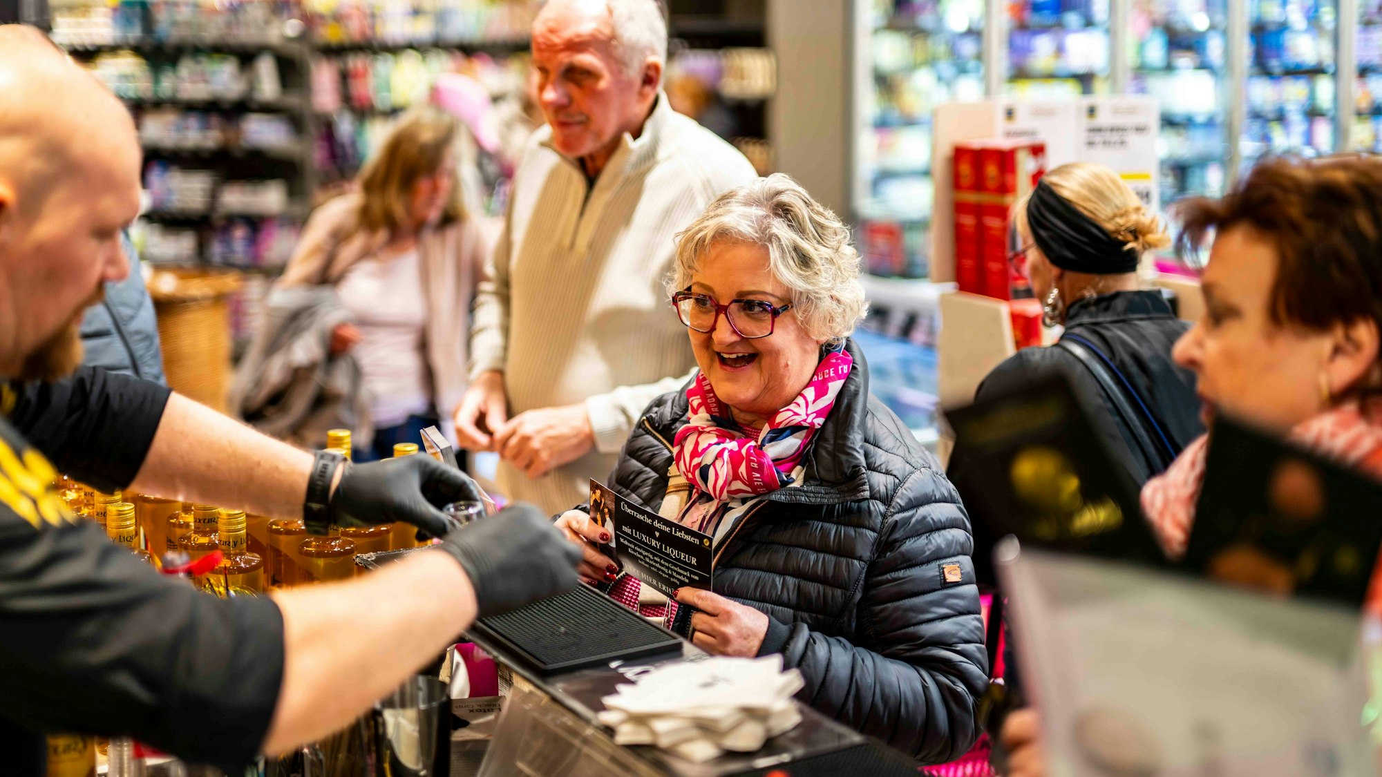 Das Bild zeigt die Seniorin während des Events. Sie steht an einer Theke und wartet auf einen Liquör.