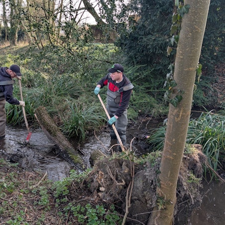Das Foto zeigt zwei Bauarbeiter in Wasserschutzhosen und mit Spaten im Bachlauf des Gleueler Bachs.