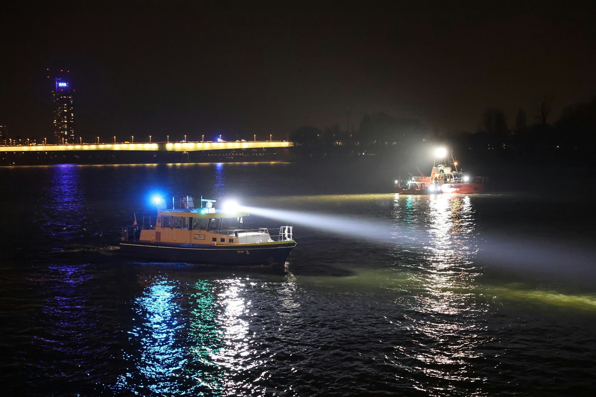 07.03.2026, Nordrhein-Westfalen, Köln: Nachdem ein PKW in in den Rhein gestürzt war, suchen ein Polizeiboot (l) und ein weiteres Boot das Wasser ab. Das Auto war bei einem Unfall am Kölner Rheinufer mitsamt den Insassen, einem jungen Mann und einer jungen Frau im Fluss gelandet. Der Mann wurde lebensgefährlich verletzt. Foto: Jan Ohmen/-/dpa +++ dpa-Bildfunk +++