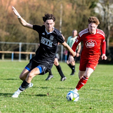 Zwei Fußballer im Kampf um den Ball. Im Hintergrund ruft ein dritter Fußballer etwas.