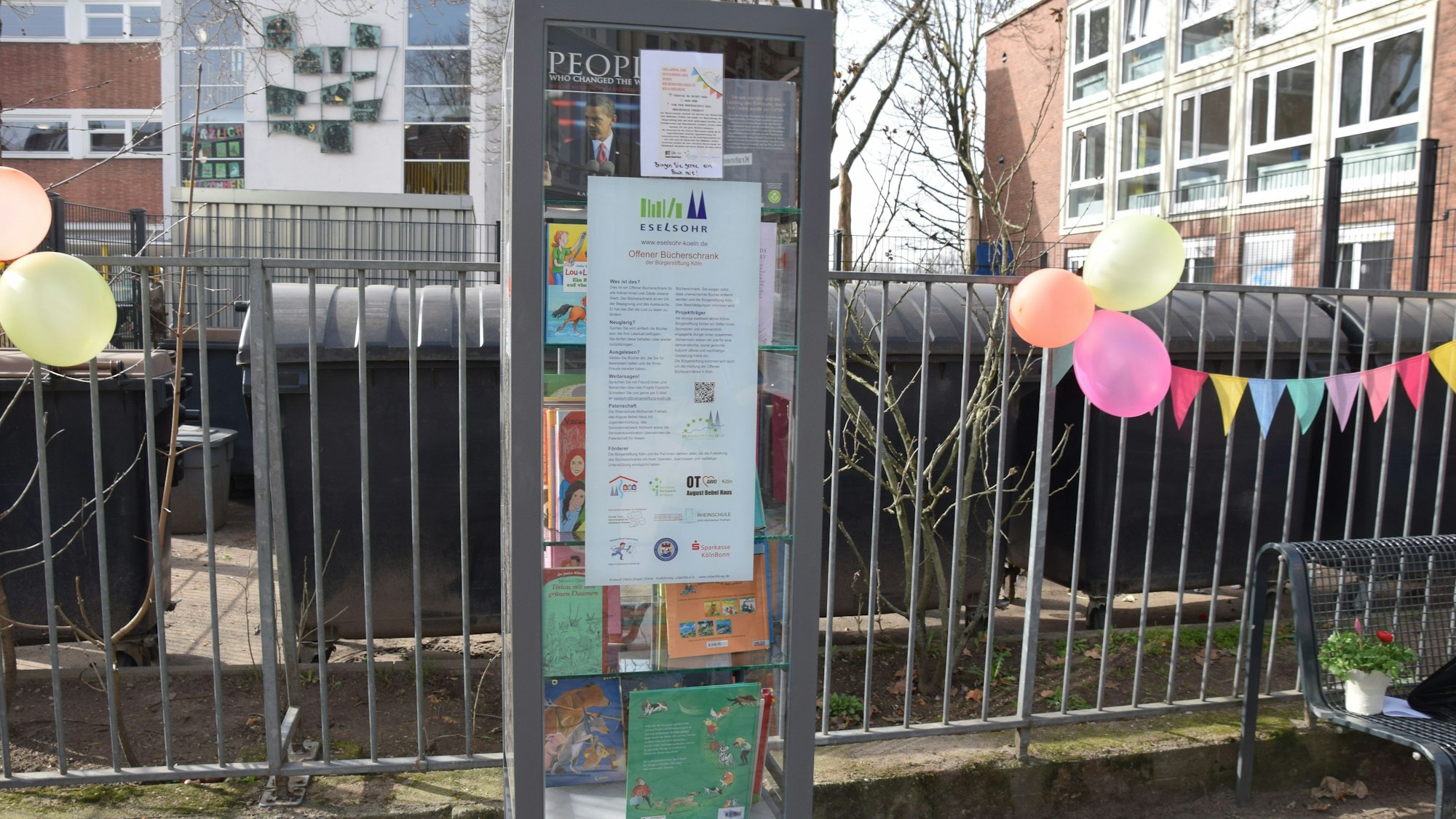 Ein Bücherschrank steht vor dem Gitterzaun, hinter dem ein Schulgelände liegt.