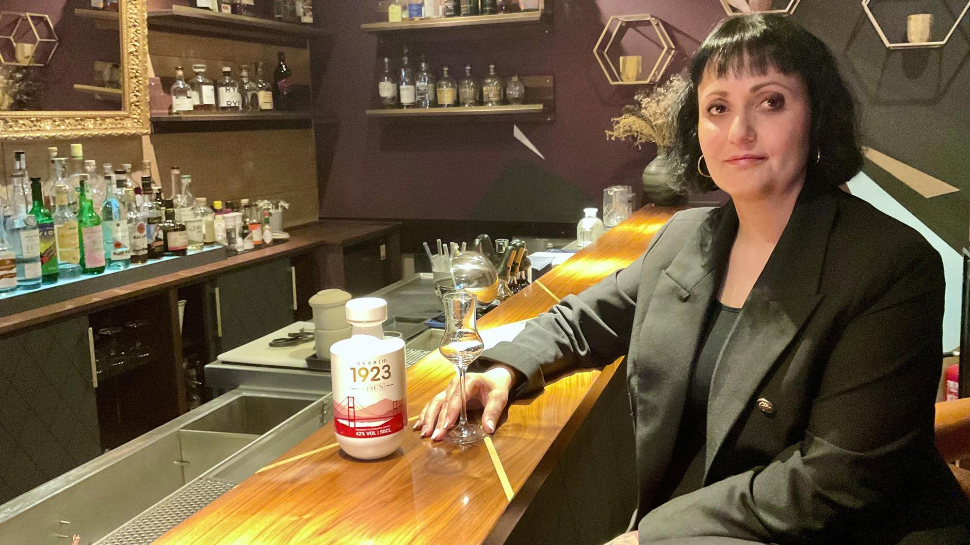 Eine Frau sitzt an einem Tressen, sie hält ein Glas, daneben steht eine Flasche mit weißem Etikett und roter Schrift.