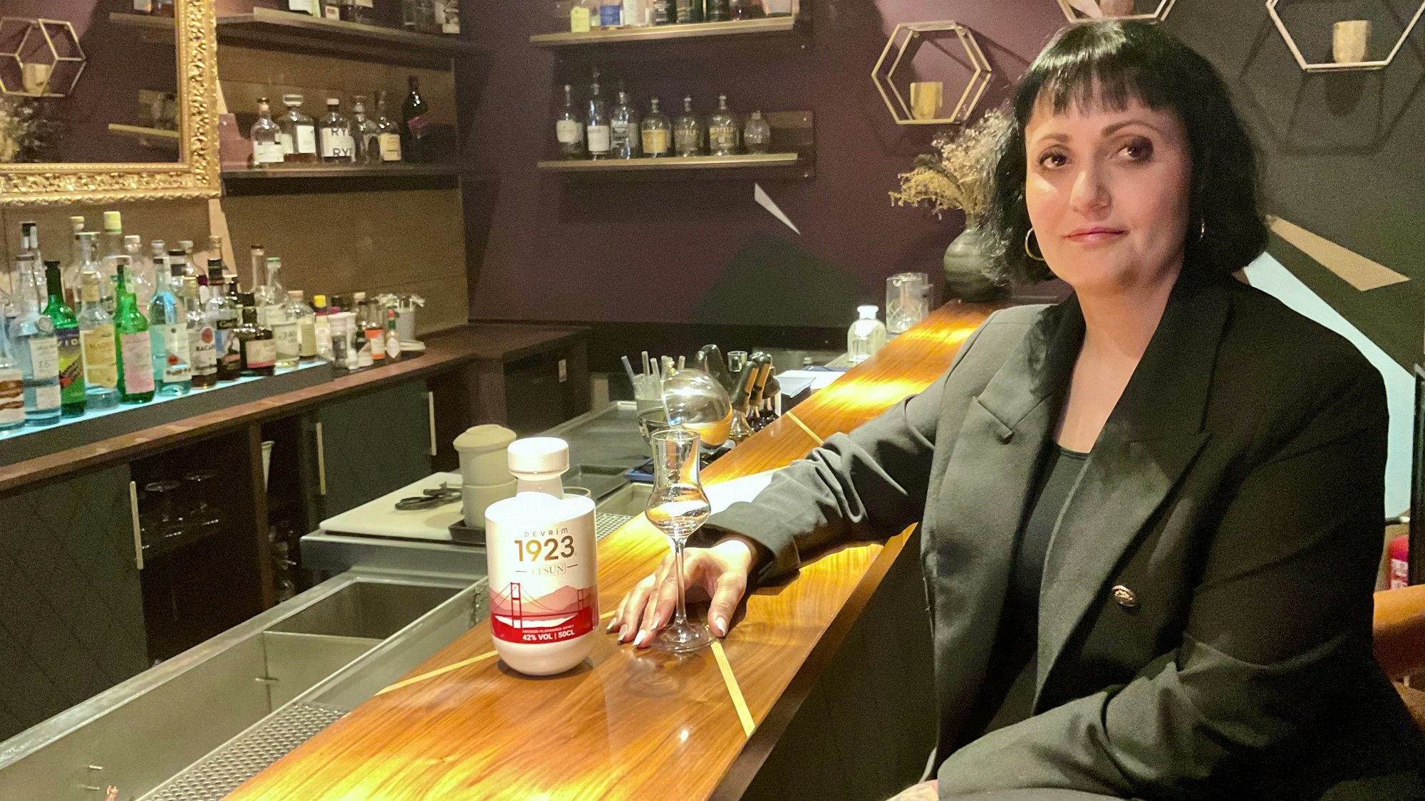 Eine Frau sitzt an einem Tressen, sie hält ein Glas, daneben steht eine Flasche mit weißem Etikett und roter Schrift.