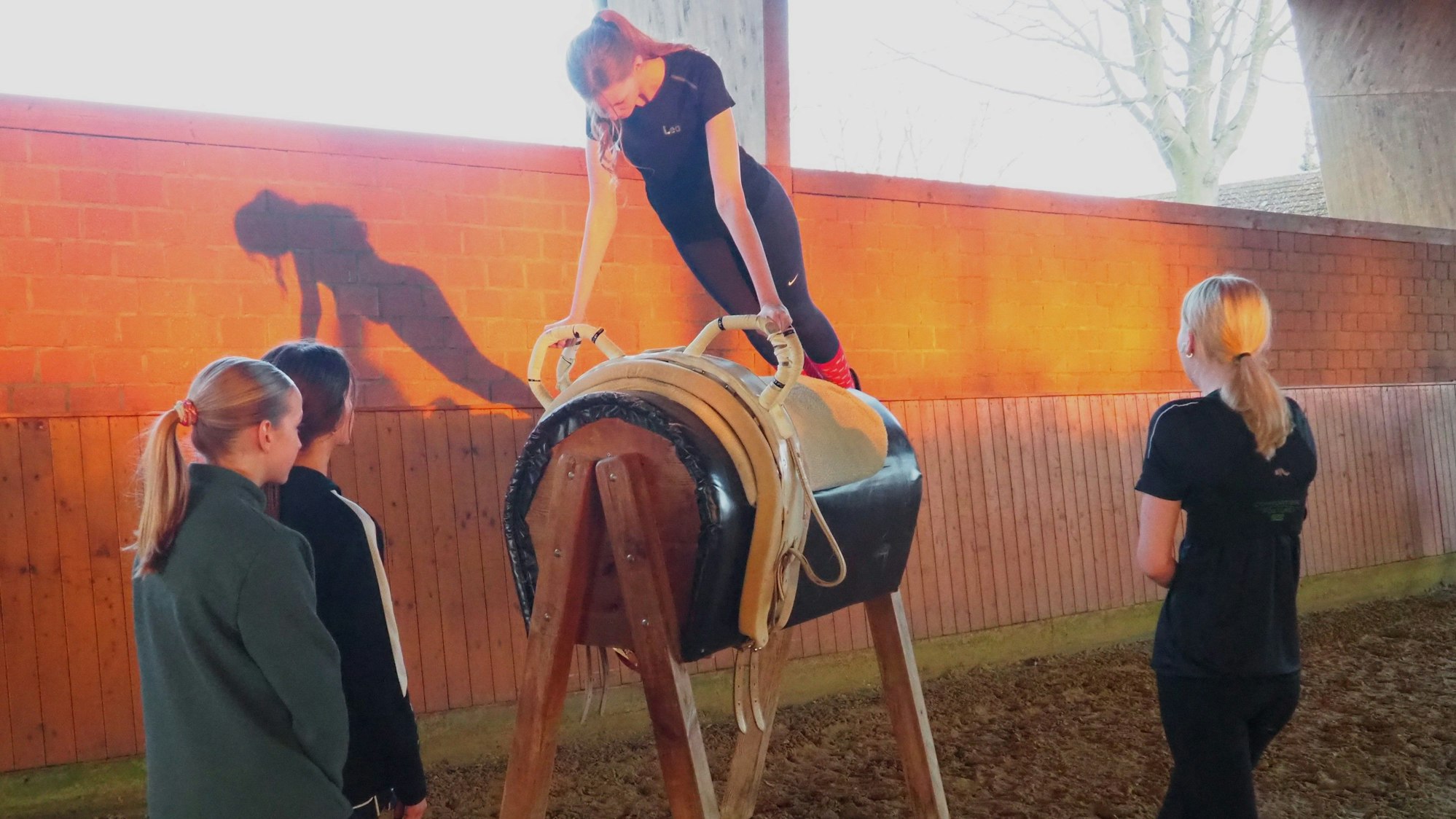 Ein Mädchen führt eine Übung auf einem Holzpferd vor, drei schauen zu.